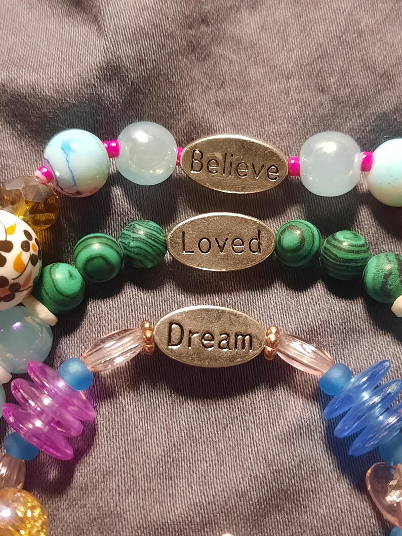 Puede incluir: Tres pulseras de cuentas con palabras inspiradoras. La pulsera superior tiene cuentas azules claras y blancas con la palabra "Believe". La pulsera del medio tiene cuentas verdes con la palabra "Loved". La pulsera inferior tiene cuentas azules y moradas con la palabra "Dream".