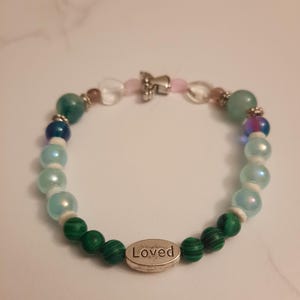 Puede incluir: Pulsera de cuentas coloridas con cuentas verdes, azules e iridiscentes. Cuenta con un dije plateado con la palabra "Loved". La pulsera est&aacute; ensartada en un cord&oacute;n el&aacute;stico transparente.