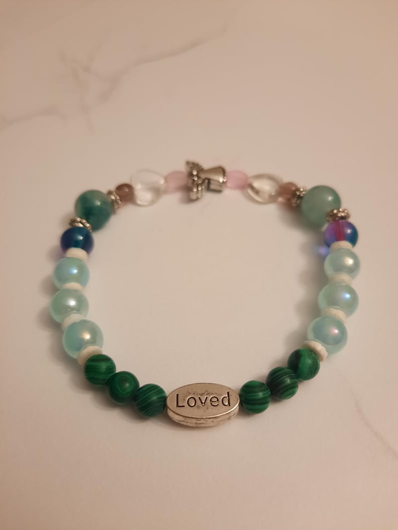 Puede incluir: Pulsera de cuentas coloridas con cuentas verdes, azules e iridiscentes. Cuenta con un dije plateado con la palabra "Loved". La pulsera est&aacute; ensartada en un cord&oacute;n el&aacute;stico transparente.