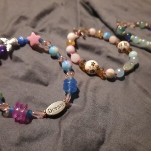 Puede incluir: Tres pulseras de cuentas con varias cuentas de colores. Una pulsera presenta cuentas en forma de estrella rosa, cuentas azules y transparentes y un dije plateado. Otra pulsera tiene la palabra "Dream" en una cuenta ovalada plateada.