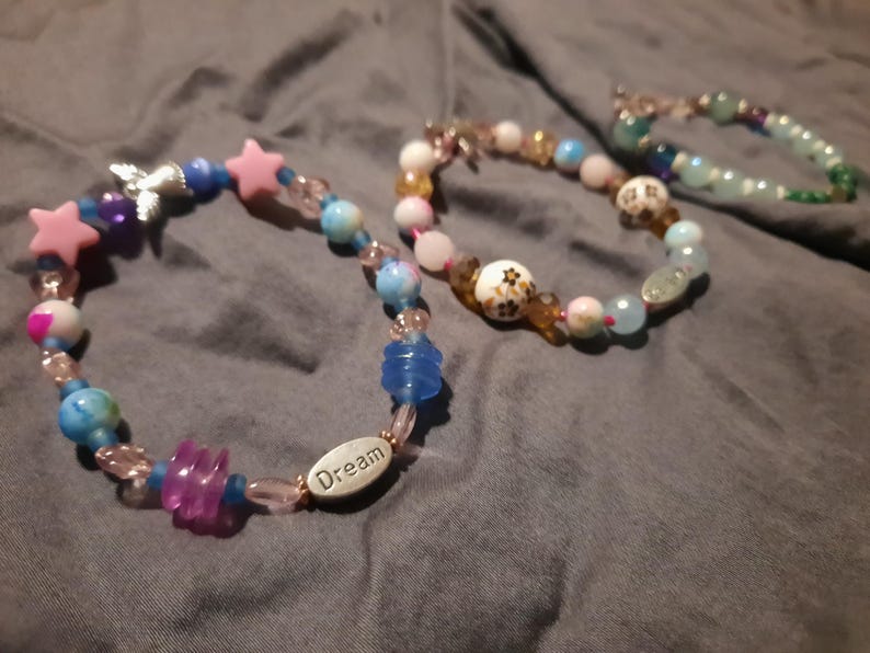 Puede incluir: Tres pulseras de cuentas con varias cuentas de colores. Una pulsera presenta cuentas en forma de estrella rosa, cuentas azules y transparentes y un dije plateado. Otra pulsera tiene la palabra "Dream" en una cuenta ovalada plateada.