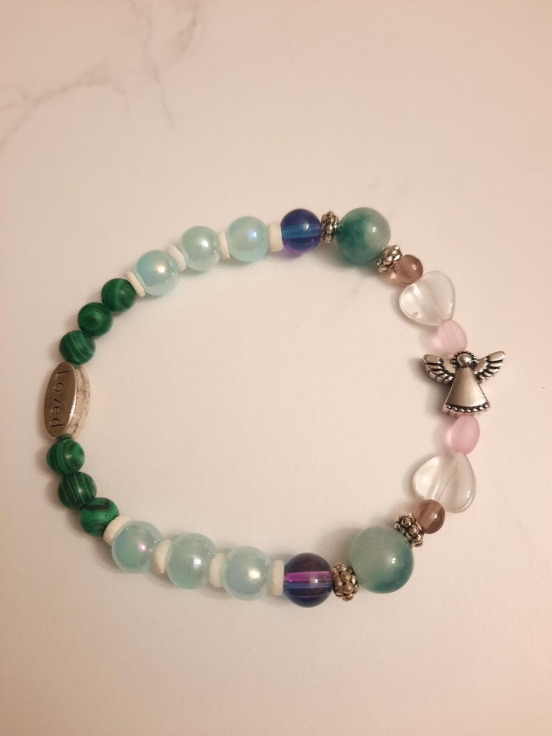 Puede incluir: Una pulsera de cuentas con cuentas verdes de malaquita, azules iridiscentes y rosas en forma de coraz&oacute;n. Tambi&eacute;n hay un dije de &aacute;ngel plateado y un dije plateado con la palabra "Loved".