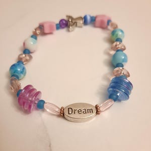 May include: A beaded bracelet with a variety of colorful beads in shades of blue, pink, and clear. The bracelet includes a silver charm and a silver oval charm with the word "Dream" engraved on it.
