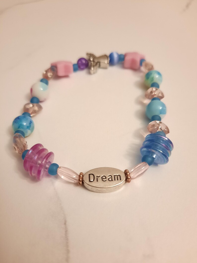 May include: A beaded bracelet with a variety of colorful beads in shades of blue, pink, and clear. The bracelet includes a silver charm and a silver oval charm with the word "Dream" engraved on it.