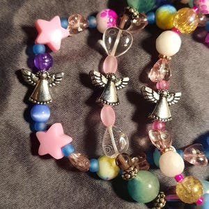 May include: A beaded bracelet featuring a variety of colorful beads, including pink stars, clear heart-shaped beads, and silver angel charms. The beads are in shades of pink, blue, purple, yellow, and green. The bracelet is on a gray surface.