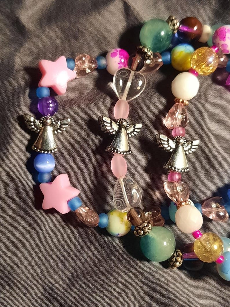 May include: A beaded bracelet featuring a variety of colorful beads, including pink stars, clear heart-shaped beads, and silver angel charms. The beads are in shades of pink, blue, purple, yellow, and green. The bracelet is on a gray surface.