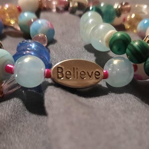 May include: Close-up of beaded bracelets with various colorful beads. One bracelet has the word "Dream" on an oval charm, another has "Believe", and a third has "Loved". The beads are in shades of blue, green, pink, and white.