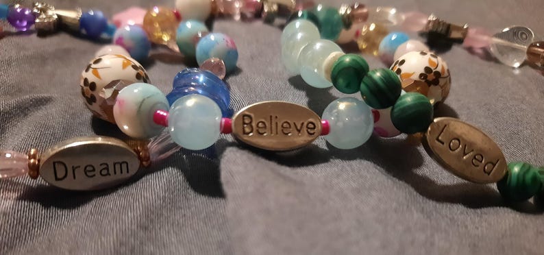 May include: Close-up of beaded bracelets with various colorful beads. One bracelet has the word "Dream" on an oval charm, another has "Believe", and a third has "Loved". The beads are in shades of blue, green, pink, and white.