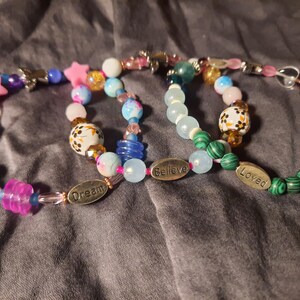 Puede incluir: Tres pulseras de cuentas de colores. Las pulseras presentan una variedad de cuentas, incluyendo estrellas rosas, cuentas azules y blancas estampadas y cuentas transparentes y de colores. Una pulsera tiene un dije que dice "Dream", otra "Believe" y la tercera "Loved".