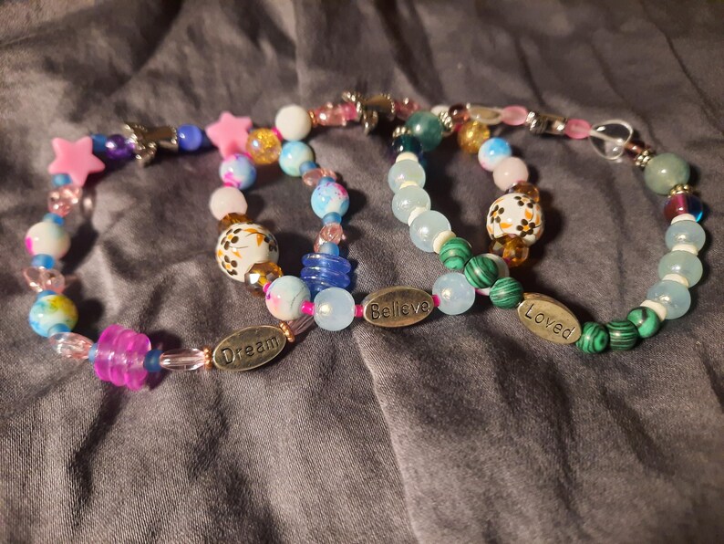 Puede incluir: Tres pulseras de cuentas de colores. Las pulseras presentan una variedad de cuentas, incluyendo estrellas rosas, cuentas azules y blancas estampadas y cuentas transparentes y de colores. Una pulsera tiene un dije que dice "Dream", otra "Believe" y la tercera "Loved".