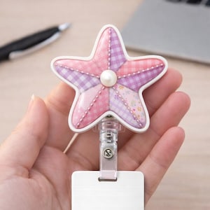 May include: A retractable badge holder with a pink and purple patchwork starfish design, a white border, and a pearl accent. The holder has a clear plastic clip and a blank white rectangular badge.
