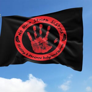 May include: Black flag with a red handprint inside a red circle, with the text "No More Stolen Sisters Missing & Murdered Indigenous Women." The flag is waving in the wind against a blue sky.