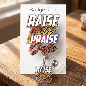 May include: A badge reel with the words "RAISE HELL PRAISE DALE" in a layered, glittery design. The text is in white, orange, yellow, and blue, with a silver glitter background. The reel is attached to a clear plastic clip.