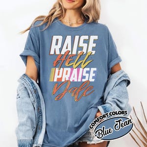 May include: Blue Comfort Colors t-shirt with the words "RAISE HELL PRAISE DALE" in a retro font. The shirt has a vintage look and feel, with a slightly faded appearance. The text is in white, yellow, orange, and blue.