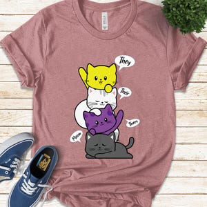 May include: A dusty rose t-shirt featuring a graphic of four cartoon cats in yellow, white, purple, and gray. Speech bubbles above the cats read "They", "Their", and "Them". A speech bubble says "Meow".