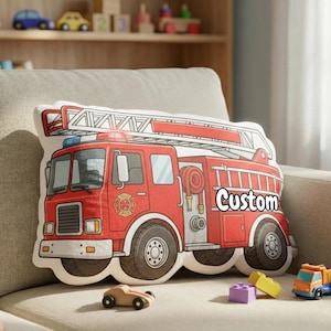 May include: A decorative pillow shaped like a red fire truck with a ladder on top. The word "Custom" is printed on the side of the truck. The pillow is on a beige couch with toy cars and blocks.