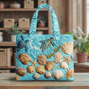 Puede incluir: Bolso tote turquesa con bordados de tortugas marinas y conchas. El bolso tiene un diseño acolchado con patrones de olas y el texto "Your Name" en escritura cursiva. Las asas son turquesas a juego con un estampado floral blanco.