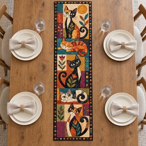 May include: A rectangular fabric table runner featuring a patchwork design of five distinct panels, each depicting stylized cats in various poses, surrounded by a black border with colorful dots, set on a wooden table with four place settings.