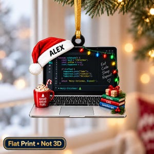 May include: A flat hanging ornament shaped like a laptop with a red Santa hat labeled ALEX, a red mug with GOOD IDEAS FUEL, and a stack of books labeled HTML, CSS, and JavaScript. The screen displays code and the text Eat Code Sleep Repeat.
