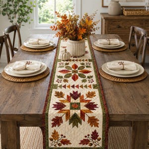 May include: A quilted table runner featuring a repeating pattern of autumn leaves in shades of orange, red, brown, and green on a cream background. The runner is centered on a wooden dining table with white plates, napkins, and a vase of fall foliage.