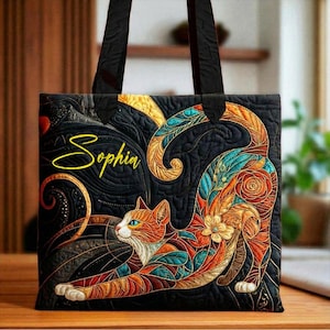 May include: Black tote bag featuring a colorful cat design with orange, teal, and gold accents. The cat is stretching, with the name "Sophia" in yellow script. The bag has black handles and a quilted texture.