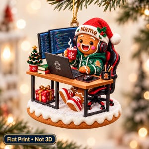 May include: A gingerbread person ornament wearing a green hoodie and Santa hat, sitting at a desk with a laptop, two monitors displaying code, a mug, and a small Christmas tree. A gift box sits below the desk on a snowy base. Text reads Name.