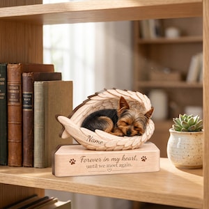 May include: A wooden pet memorial featuring a sleeping Yorkshire Terrier nestled within carved angel wings. The base is engraved with the text 'Forever in my heart, until we meet again.' and includes a space for a name on the wing.