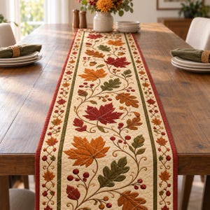May include: A rectangular quilted table runner featuring a cream-colored center panel decorated with embroidered autumn leaves in shades of red, orange, and green, accented by small berries and acorns, bordered by dark red and green fabric trim.