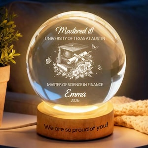 Puede incluir: Una esfera de graduación de vidrio transparente con una base de madera, iluminada desde el interior. La esfera está grabada con las palabras "Mastered it!" y "University of Texas at Austin", junto con un birrete, un diploma, flores, el nombre "Emma" y el año "2026". La base dice "We are so proud of you!"