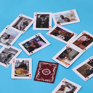 May include: A collection of custom playing cards featuring various personal photographs of a couple, scattered across a blue surface. One card displays a dark red back with white ornate filigree borders and the text E&J 04.22.2024 in the center.