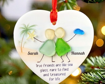 May include: A heart-shaped white ceramic ornament featuring two figures made of sea glass, one light green and one dark green. The design includes a yellow starfish, a blue cloud, palm trees, the names Sarah and Hannah, and the text True friends are like sea glass, rare to find and treasured for life, suspended by a red ribbon.