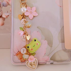 May include: Clear phone case with a floral design and a gold-colored charm. The charm includes a star, a pearl, pink flowers, a green frog, and the letters A, M, and Y. The case has a pink border.