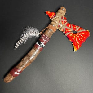 May include: A decorative tomahawk featuring a bright red and orange stone-like blade secured to a wooden handle with tan leather lacing. The handle is wrapped with dark red, black, and white leather strips and adorned with a grey and white striped feather.