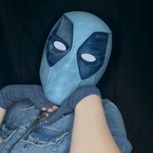 May include: A light blue full-head mask with dark blue eye patches and white oval eye cutouts. The mask features a textured surface and is worn with dark blue fingerless gloves and a blue denim jacket with a visible metal zipper down the front.