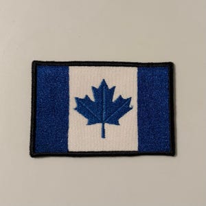 May include: A rectangular embroidered patch featuring a blue and white triband design. The center white panel displays a blue maple leaf, while the two side panels are solid blue. The entire patch is bordered by a thick, black stitched edge.