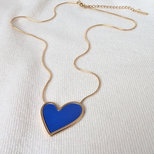 Könnte beinhalten: Eine goldfarbene Halskette mit einem blauen Herzanhänger. Das Herz ist goldfarben umrandet und mit einem leuchtenden Blau gefüllt. Die Halskette wird auf einer weißen, strukturierten Oberfläche präsentiert.