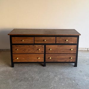 May include: A wooden dresser with a dark brown top and drawer fronts. The drawers have brass knobs and are set in a black frame. The dresser has seven drawers and is a medium-sized piece of furniture.