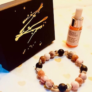May include: A bracelet featuring a mix of pink, brown, and black beads, with gold-coloured metal accents. A small bottle with a dropper and a black box with gold splatter art are also in the image. The bracelet is a fashion accessory.