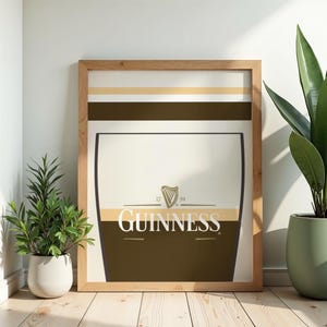 Op de afbeelding: Ingelijste Guinness-print met een pintglasontwerp, het Guinness-logo en het harpsymbool. De print heeft een lichtbruine houten lijst. De afbeelding staat tegen een witte muur, met potplanten aan weerszijden.