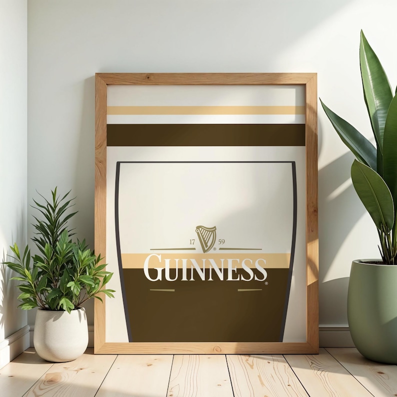 Op de afbeelding: Ingelijste Guinness-print met een pintglasontwerp, het Guinness-logo en het harpsymbool. De print heeft een lichtbruine houten lijst. De afbeelding staat tegen een witte muur, met potplanten aan weerszijden.