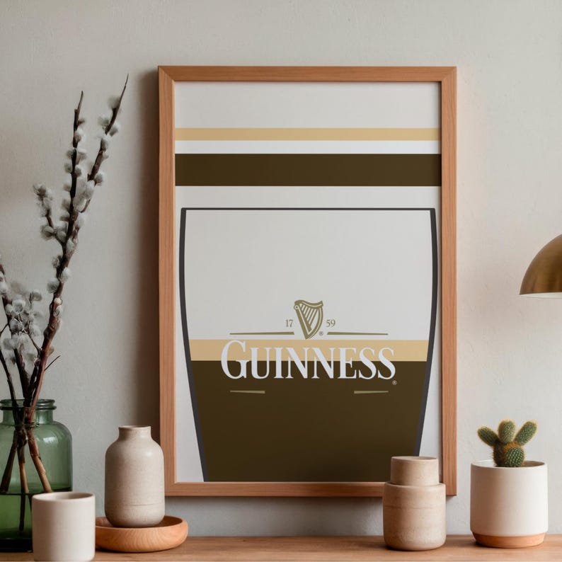 Op de afbeelding: Een ingelijste print met een gestileerd Guinness pintglasontwerp. Het kunstwerk bevat het Guinness-logo, een harpsymbool en de data 17 en 59. De print heeft een lichtbruine houten lijst en wordt gepresenteerd op een houten oppervlak.