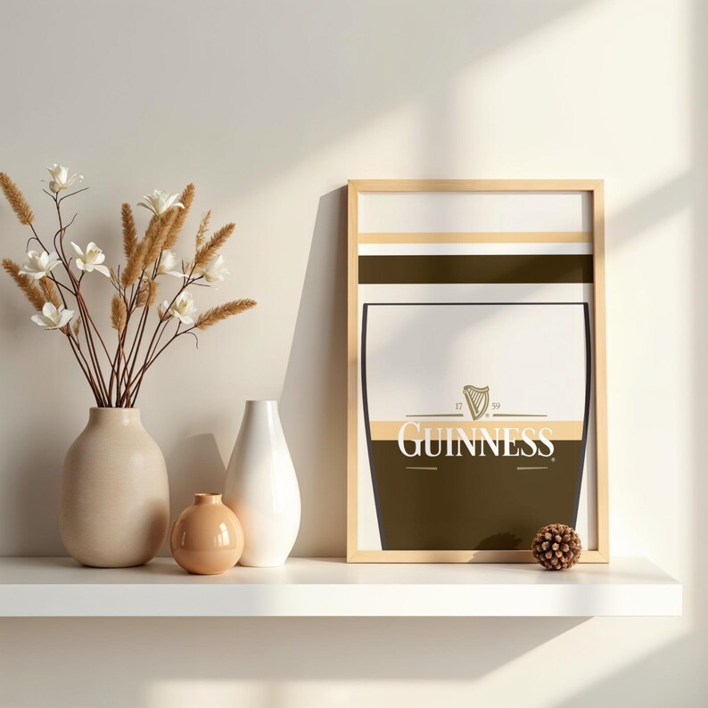 Op de afbeelding: Een ingelijste Guinness-print met een licht houten frame. De print toont een gestileerd pintglasontwerp met het Guinness-logo en de tekst. De print wordt weergegeven op een witte plank met vazen en decoratieve bloemen.