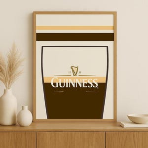 Op de afbeelding: Een ingelijste print met een gestileerd Guinness pintglas. Het ontwerp omvat een donkerbruin onderste gedeelte, een cr&egrave;mekleurig midden met het Guinness-logo en een lichtbruine en beige bovenkant. De print zit in een houten lijst.