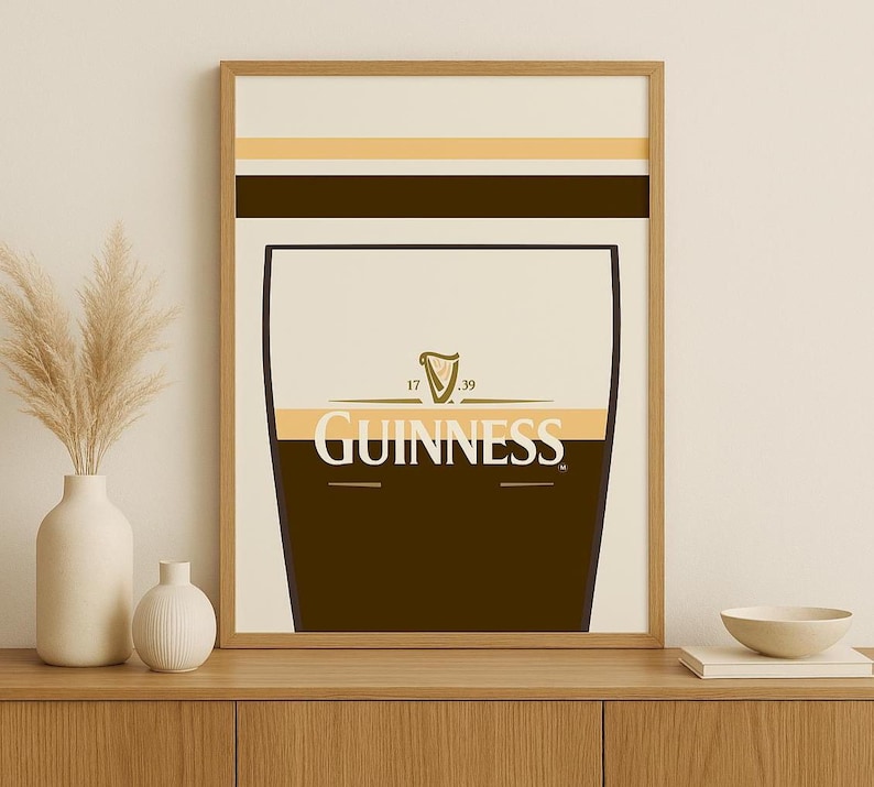 Op de afbeelding: Een ingelijste print met een gestileerd Guinness pintglas. Het ontwerp omvat een donkerbruin onderste gedeelte, een cr&egrave;mekleurig midden met het Guinness-logo en een lichtbruine en beige bovenkant. De print zit in een houten lijst.