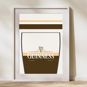 Op de afbeelding: Ingelijste kunstdruk met een gestileerde Guinness pint. Het ontwerp omvat een bruine basis, een cr&egrave;mekleurig middengedeelte en een witte bovenkant met horizontale gouden en bruine strepen. Het Guinness-logo is prominent weergegeven.