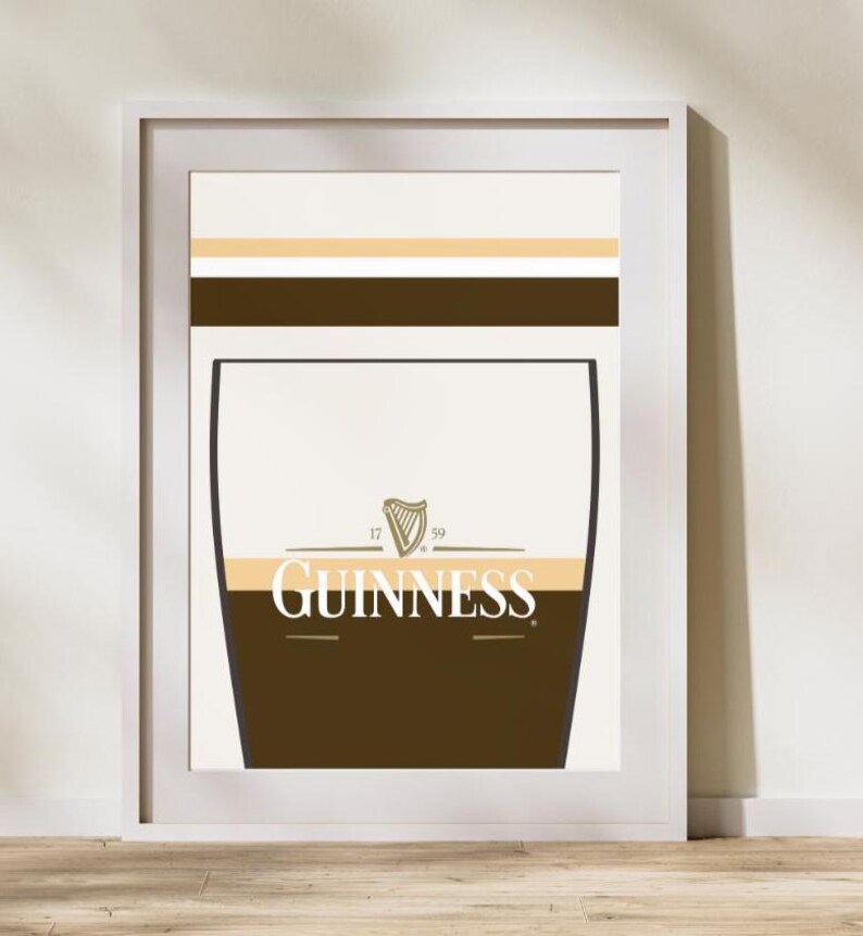 Op de afbeelding: Ingelijste kunstdruk met een gestileerde Guinness pint. Het ontwerp omvat een bruine basis, een cr&egrave;mekleurig middengedeelte en een witte bovenkant met horizontale gouden en bruine strepen. Het Guinness-logo is prominent weergegeven.