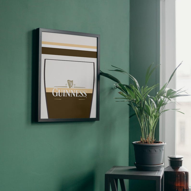 Op de afbeelding: Een ingelijste Guinness-poster met een zwarte lijst. De poster heeft een witte achtergrond met bruine en gouden designelementen. Het Guinness-logo is zichtbaar. De poster hangt aan een groene muur.