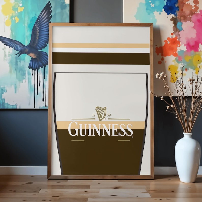 Op de afbeelding: Een ingelijste Guinness-print met een gestileerd pintglasontwerp. Het kunstwerk bevat het Guinness-logo met het harpsymbool en de tekst "GUINNESS" in het wit. De print heeft een bruin, beige en wit kleurenschema, met een houten frame.