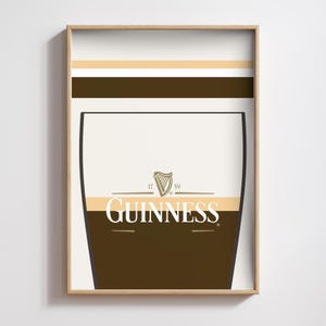 Op de afbeelding: Een ingelijste print met een gestileerd Guinness pintglas ontwerp. Het kunstwerk gebruikt wit, bruin en beige tinten, met het Guinness logo prominent weergegeven. De print zit in een lichtbruine houten lijst.