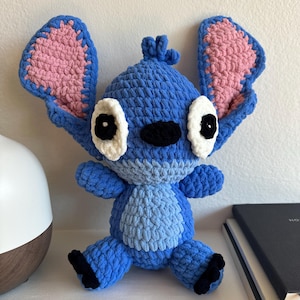 May include: A handmade crochet plush toy featuring a blue alien character with large, upright ears that have pink inner sections. The figure has white eye patches with black pupils, a black nose, and a light blue belly patch with black feet.
